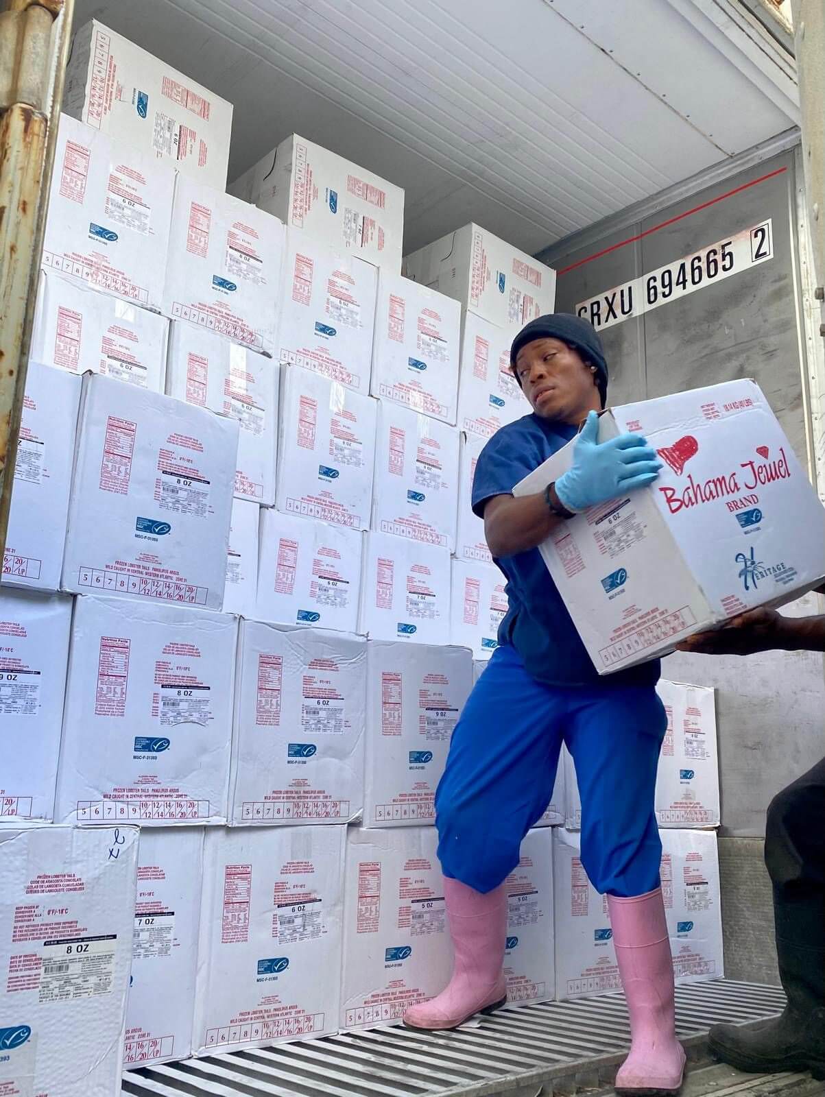 A person holding a box in front of stacked boxes in a warehouse.