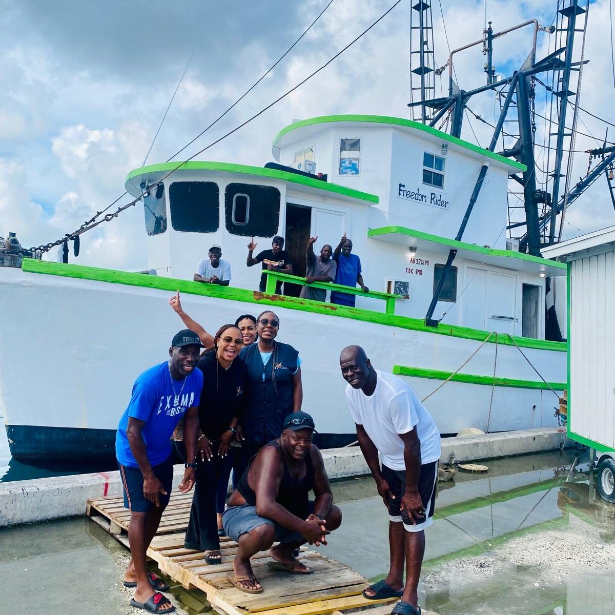 Four men posing in front of a green and white fishing boat at the dock.